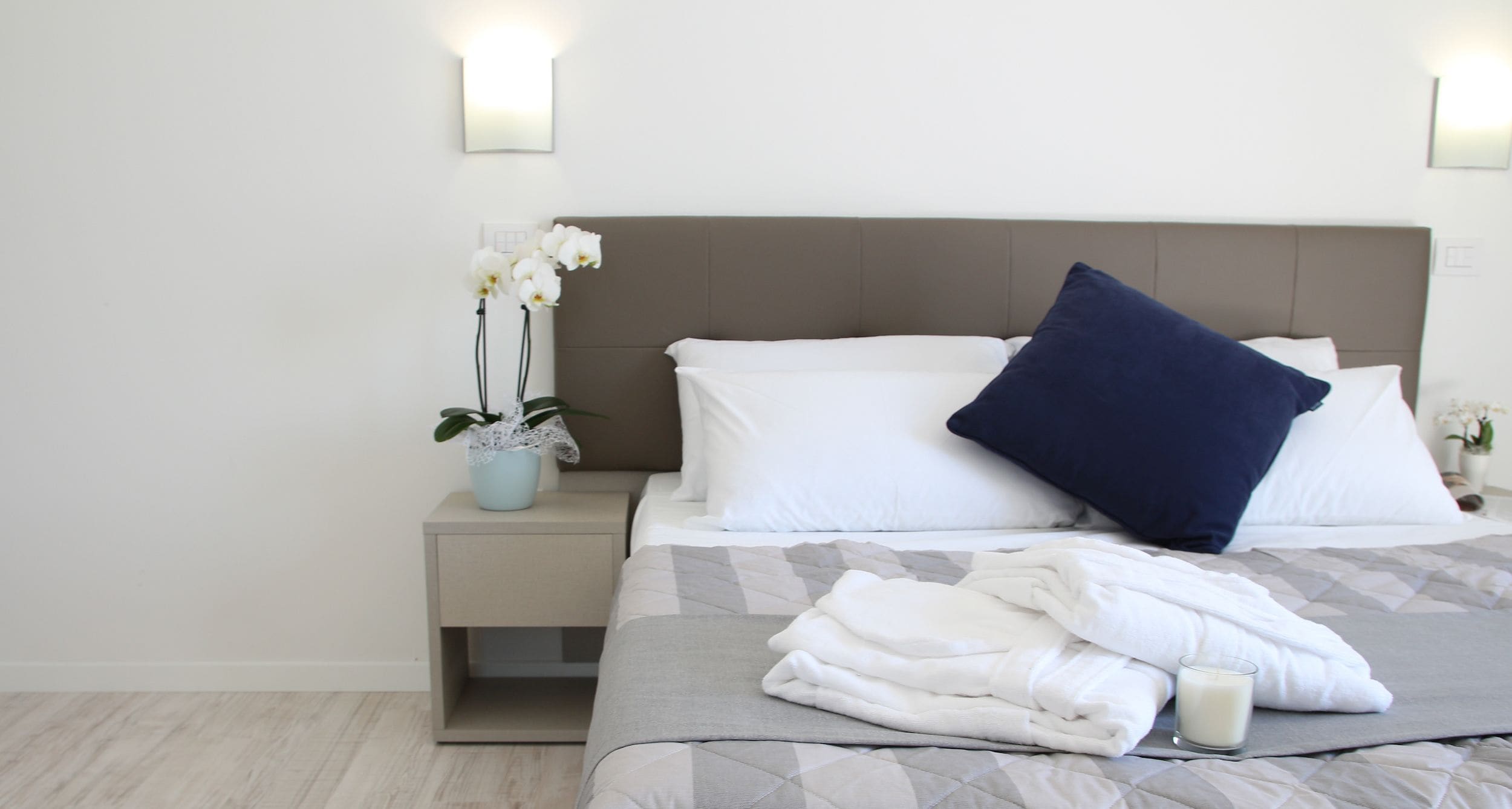 A neatly made bed with a pillow, a bedside table with a plant, and a wall-mounted light fixture.