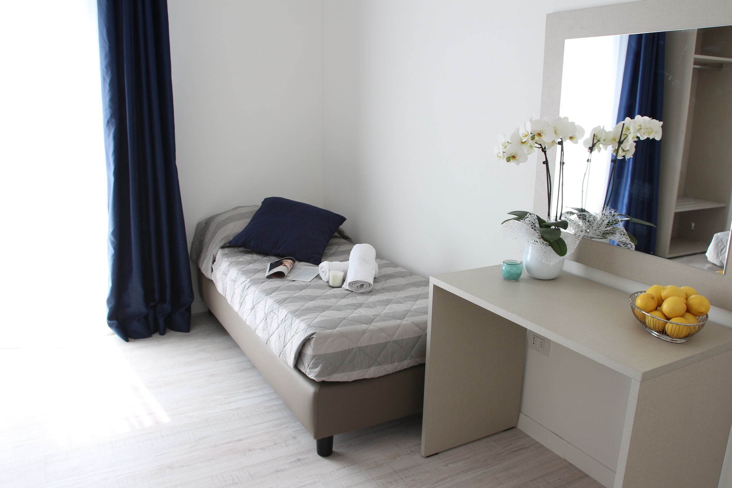 A neatly arranged bedroom with a bed, a bedside table with a bowl of fruit, a mirror, and a vase with flowers.