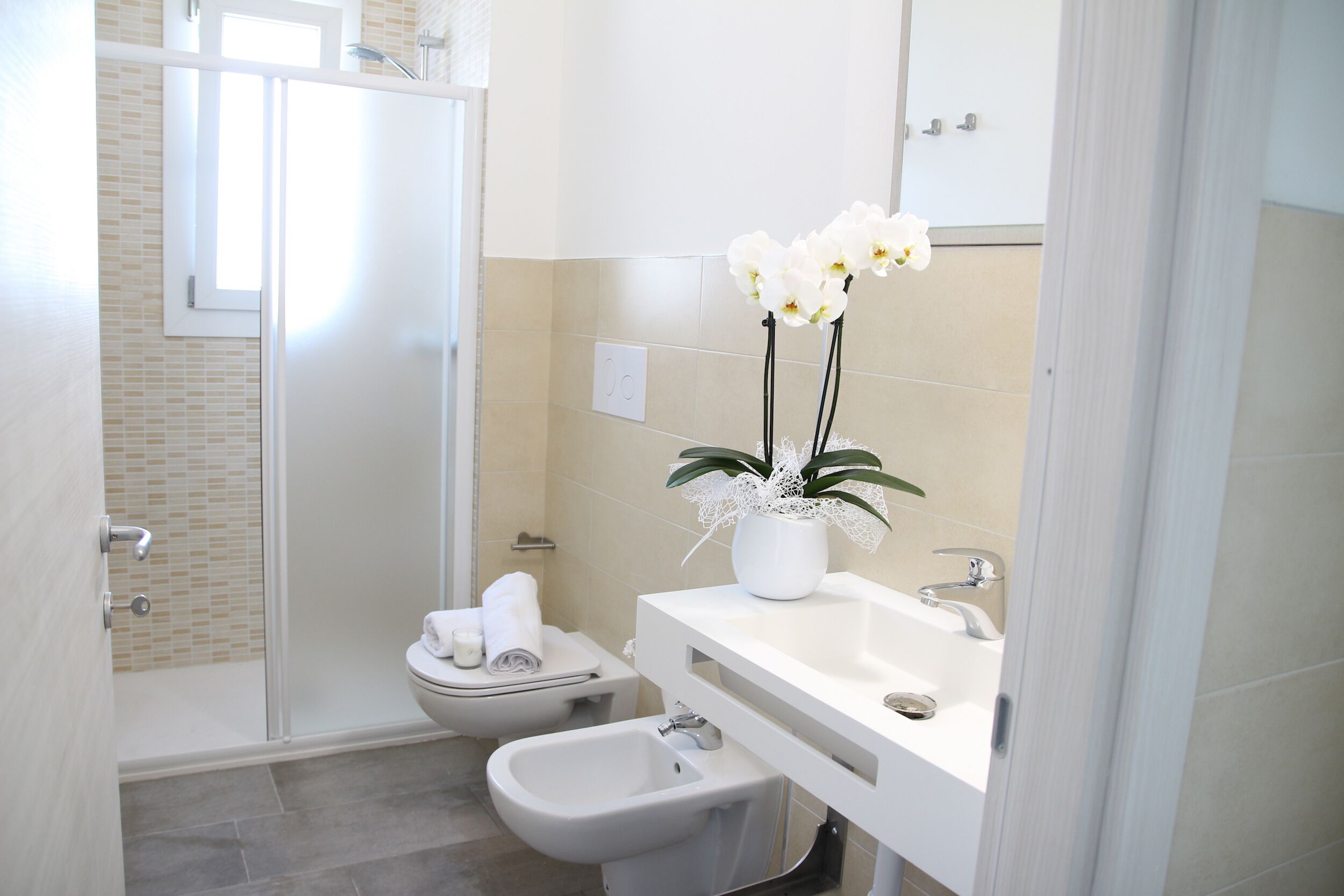 A modern bathroom with a shower, toilet, bidet, and a sink with a vase of white orchids.