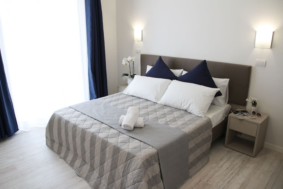 A neatly made bed with a quilted bedspread, two pillows, and a rolled towel on the bed. There is a nightstand with a vase of flowers and a magazine.