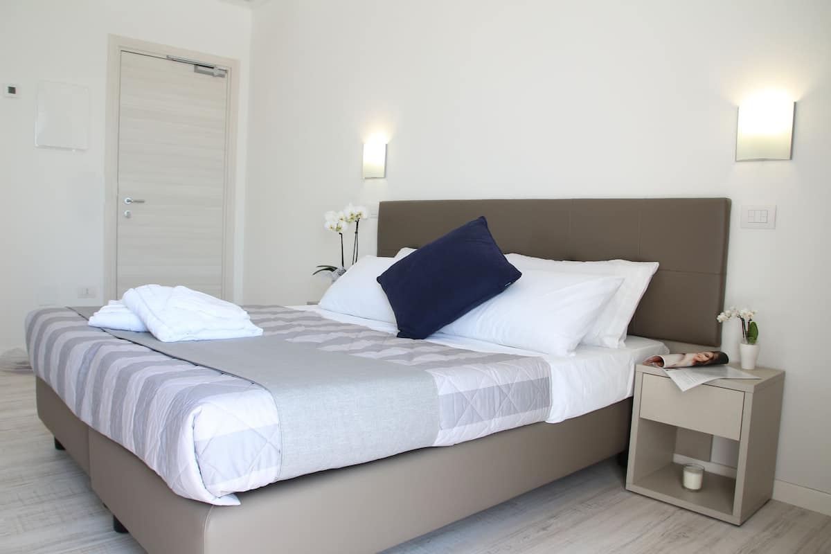 A neatly made bed with a striped blanket and pillows, a bedside table with a magazine and a small vase of flowers, and a wall-mounted light fixture.