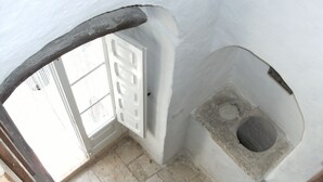 A narrow, white-walled room with a stone toilet, a door with a frosted glass panel, and a tiled floor. - Original trullo on top of a hill (Locorotondo, Bari)