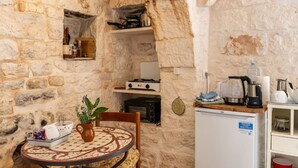 A rustic stone-walled kitchen with a small table, a chair, a potted plant, a refrigerator, and various kitchen appliances. - Original trullo on top of a hill (Locorotondo, Bari)