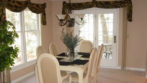 Une salle à manger avec une table dressée pour six personnes, entourée de six chaises. Un lustre est suspendu au-dessus de la table, et une plante en pot se trouve sur le côté gauche.
