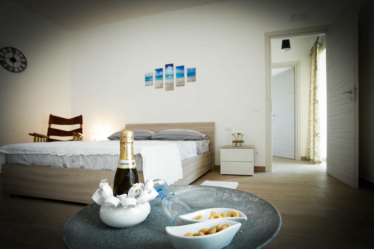 A bedroom with a bed, a bedside table, a nightstand, a wall clock, and a door leading to another room.