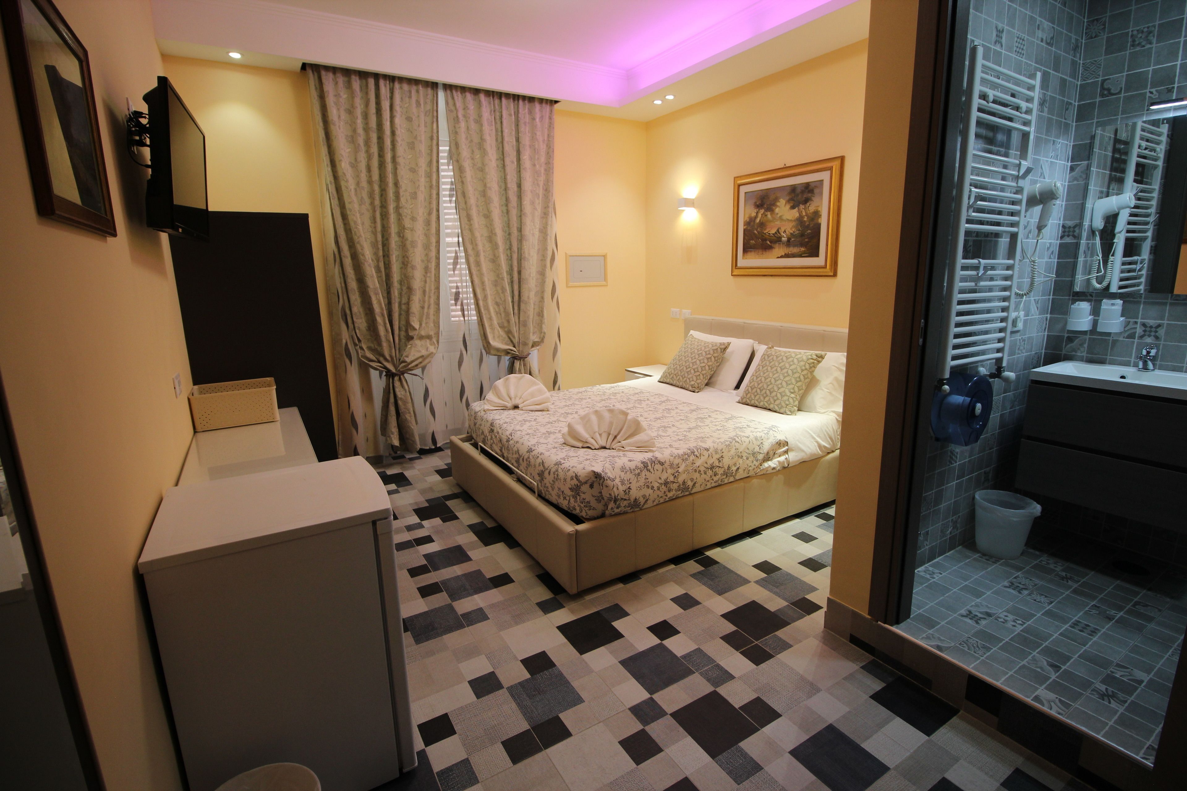 A bedroom with a bed, a bedside table, a mirror, a window with curtains, and a bathroom with a sink and towel rack.