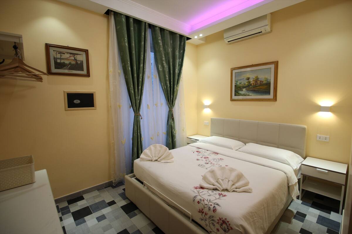 A hotel room with a bed, a bedside table, a hanging coat rack, and a wall-mounted air conditioner.