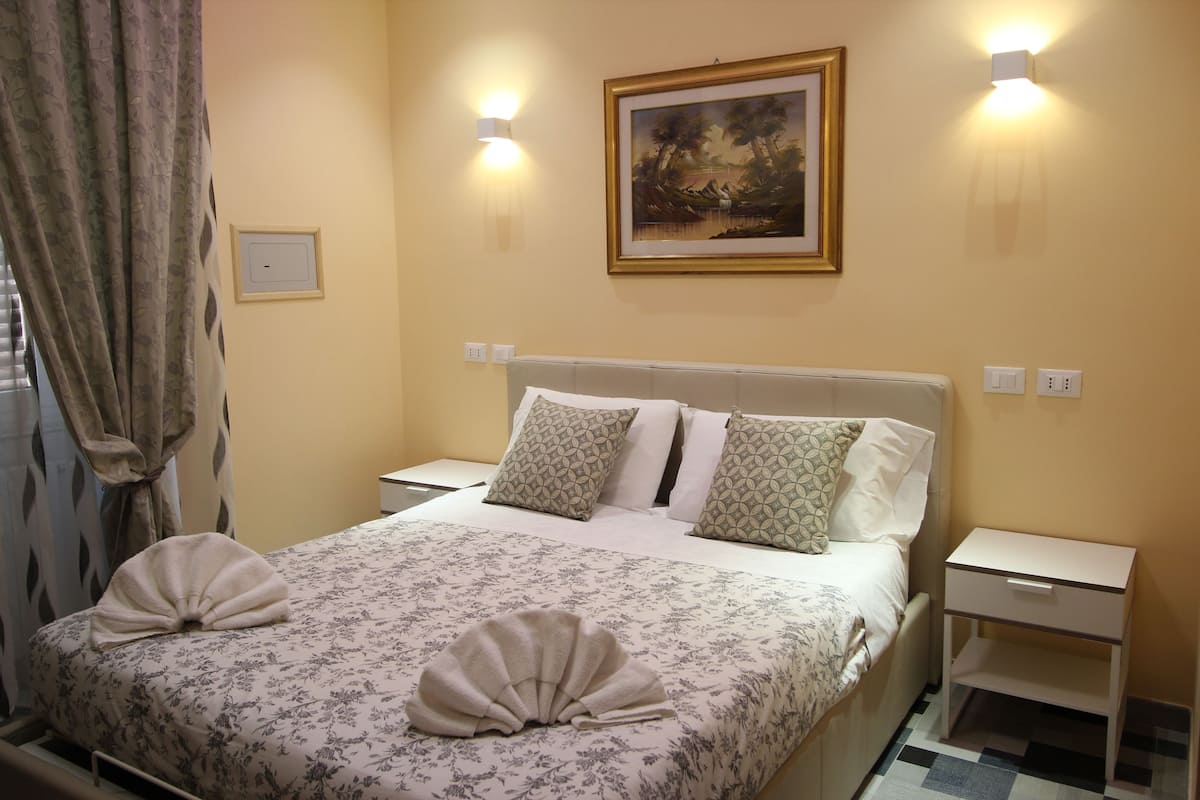 A neatly made bed with floral bedding and decorative pillows, a bedside table with a drawer, and a framed painting on the wall.