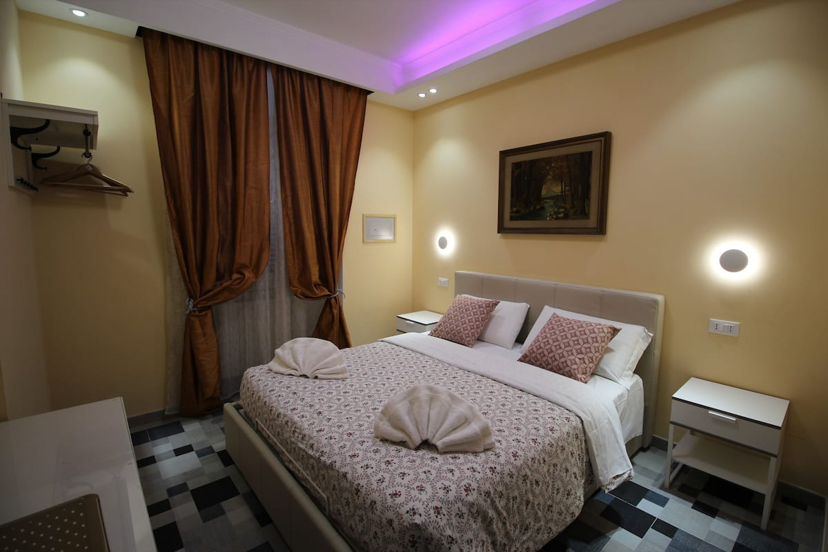 A hotel room with a bed, bedside tables, a hanging coat rack, and a painting on the wall.