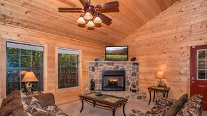 A log cabin interior with a stone fireplace, wooden walls, a ceiling fan, and a large TV on a stone mantel. - Absolutely Bearable: S/Secluded, Honeymoon, Hot Tub, Games, near Attractions! (Sevierville)