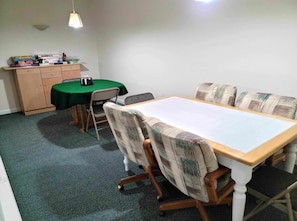 A conference room with a long table, four patterned chairs, a green tablecloth, a small table with a lamp, and a cabinet with books and a camera.