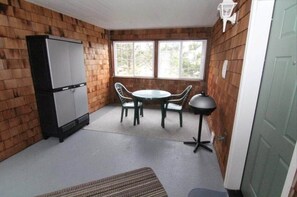 Une pièce de taille modeste avec un mur en bois, une petite table avec des chaises, un réfrigérateur et une fenêtre laissant entrer la lumière naturelle.
