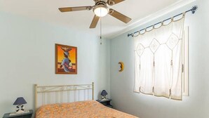 Ein Schlafzimmer mit Bett, Nachttisch, Deckenventilator, Fenster mit Vorhängen und einer Wanduhr.