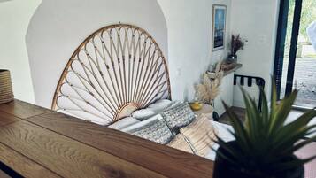 Una habitación con una mesa de madera, una silla de mimbre, una cama con almohadas, un radiador y una ventana con vistas a zonas verdes.