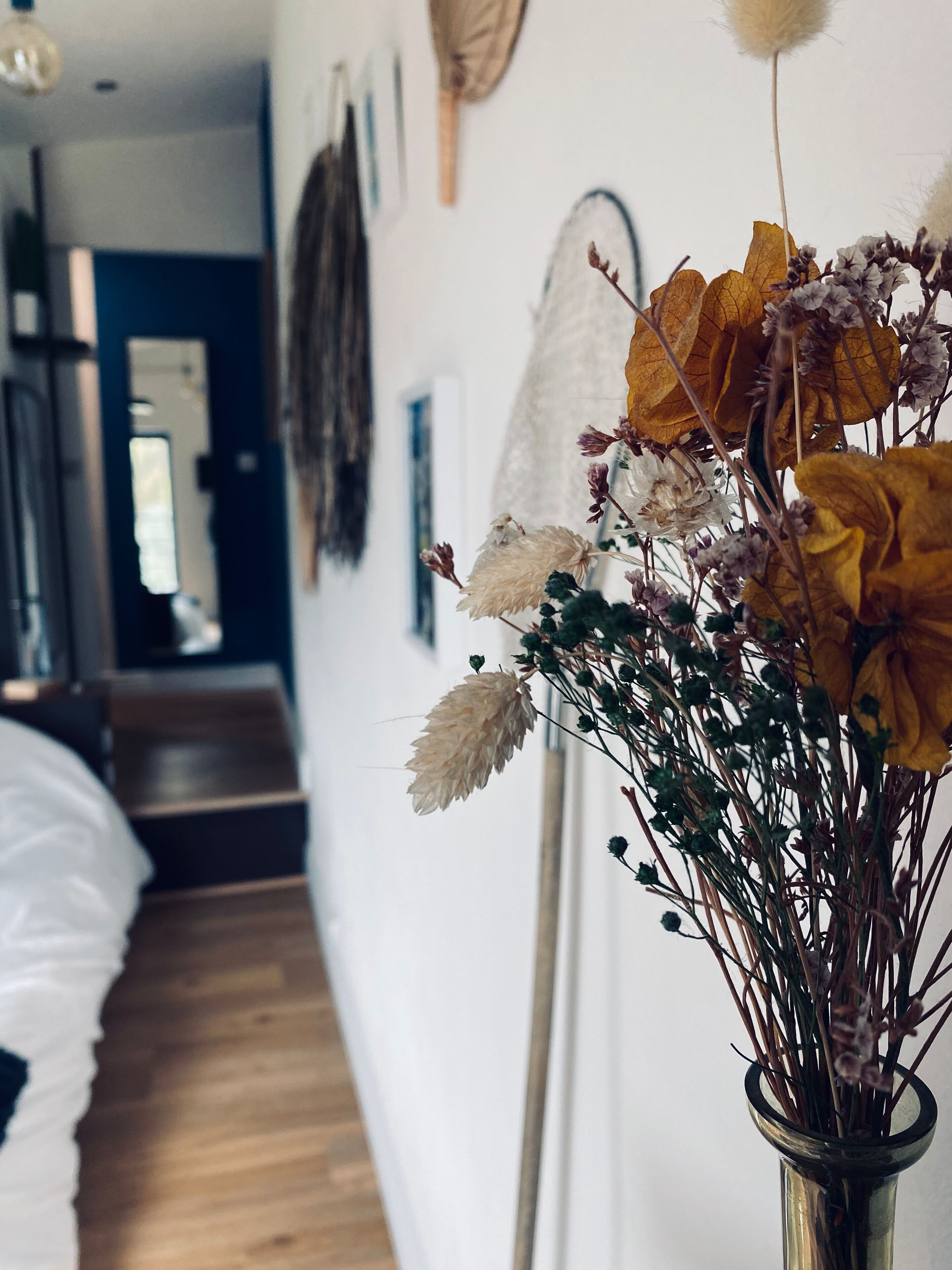 A room with a bed, a wooden floor, a vase with dried flowers, and a wall with framed pictures.