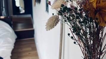 A room with a bed, a wooden floor, a vase with dried flowers, and a wall with framed pictures.
