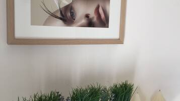 A framed photograph on a wall, a white shelf with a plant and two white pumpkins below it.