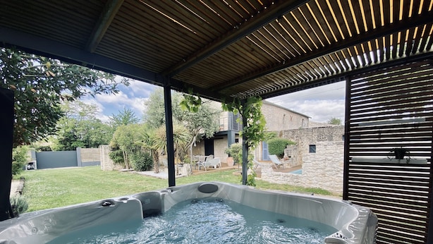 A hot tub under a wooden pergola with a view of a stone building and a grassy yard. - Ô Chambre d'hôtes (Le Gua)