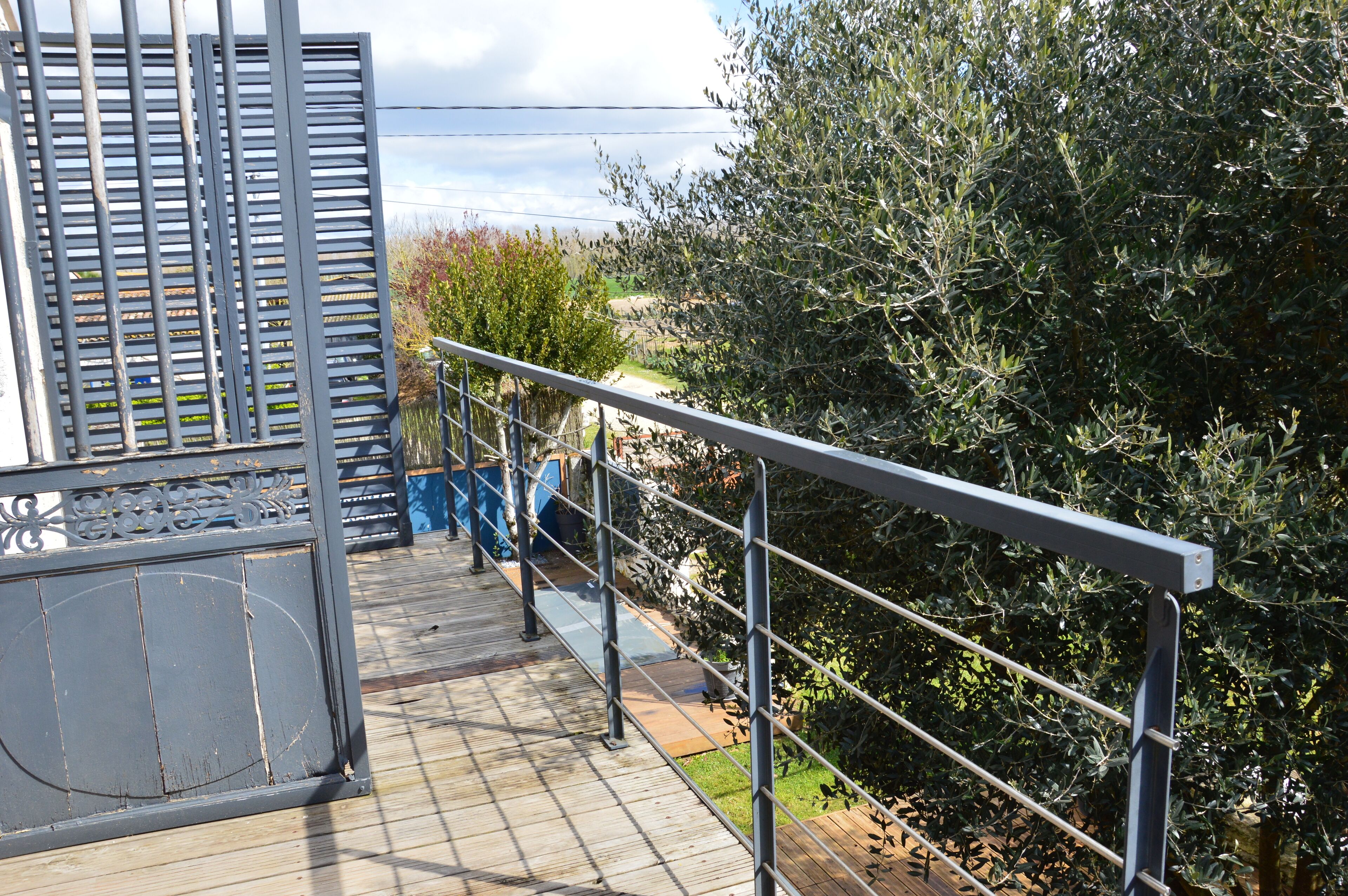 Une terrasse en bois avec des rampes en métal, donnant sur un jardin où poussent différentes plantes et offrant une vue lointaine sur un bâtiment.