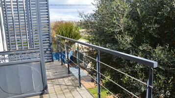 Une terrasse en bois avec des rampes en métal, donnant sur un jardin où poussent différentes plantes et offrant une vue lointaine sur un bâtiment.