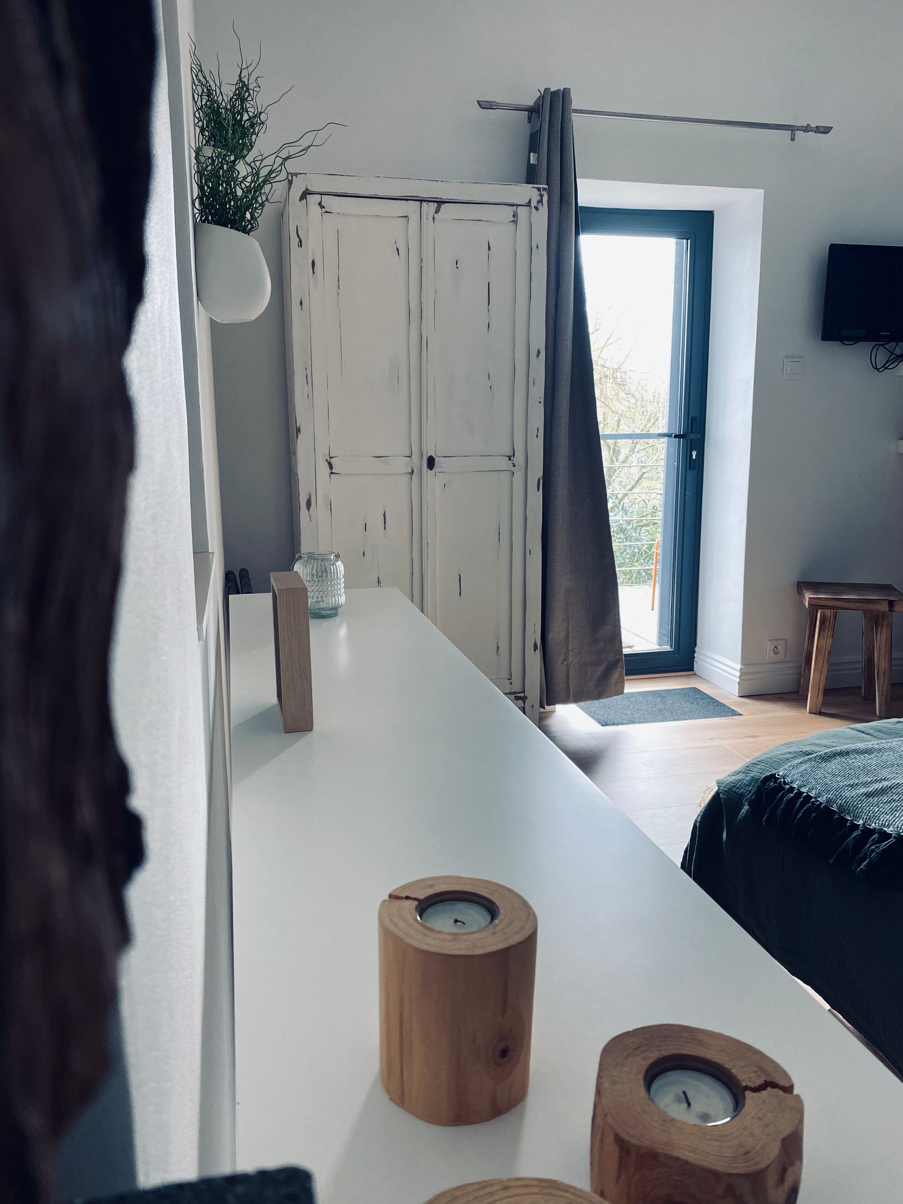 A modern bedroom with a white dresser, a wooden side table, a bed with a dark green cover, and a large window with curtains.