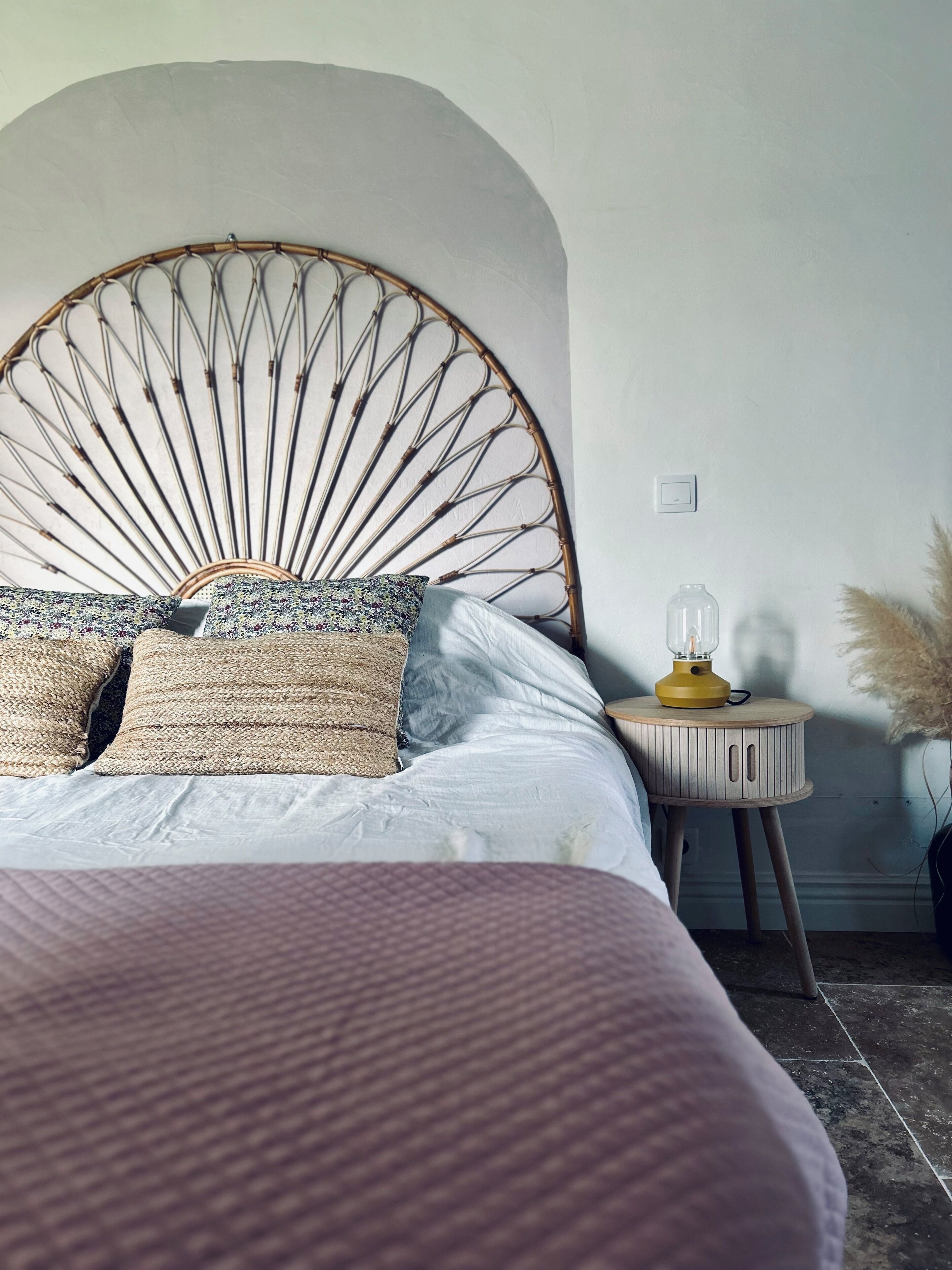 A bed with a unique bamboo headboard, a bedside table with a lamp, and a vase with pampas grass.