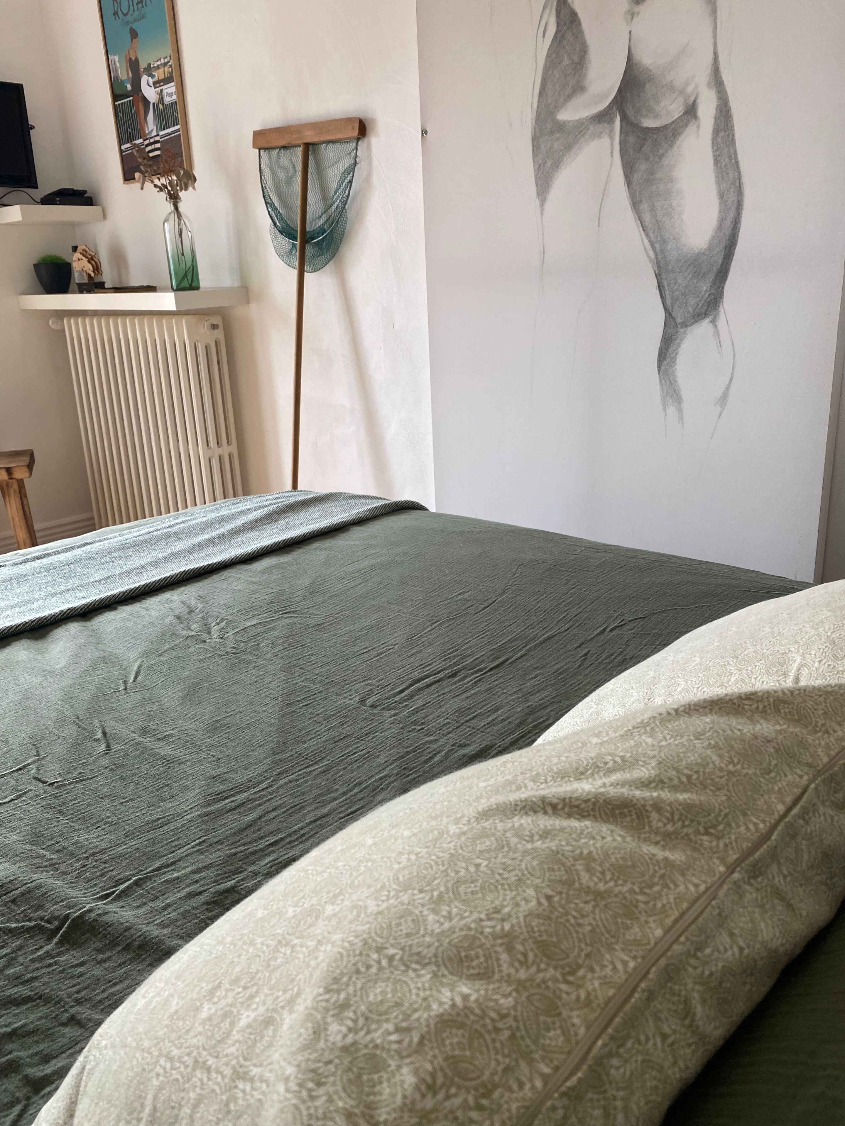 A bedroom with a bed, a radiator, a wall with a drawing, and a shelf with items.