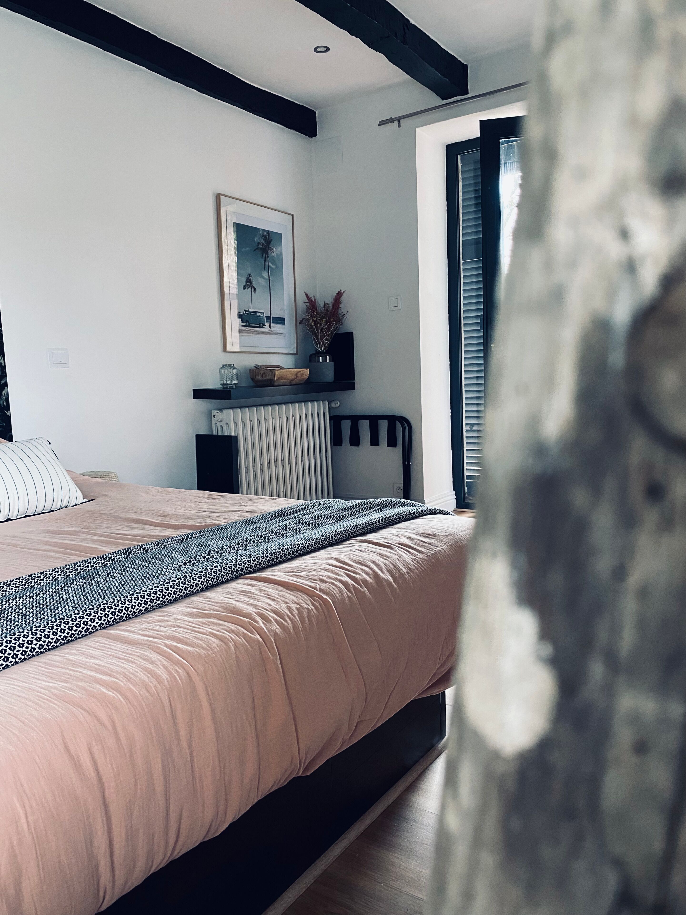 A bedroom with a bed, a radiator, a shelf with decorative items, and a window with blinds.