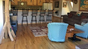 A rustic interior with a wooden ceiling, a kitchen area with wooden cabinets and bar stools, a blue patterned sofa, and a dining table.