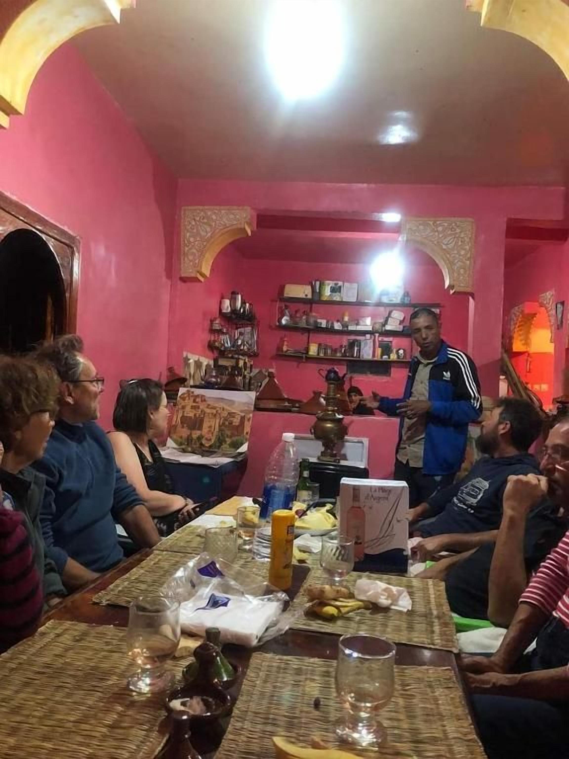 Une salle à manger où un groupe de personnes est assis autour d’une table, un serveur se tient debout, et une étagère remplie de divers objets se trouve à l’arrière-plan.