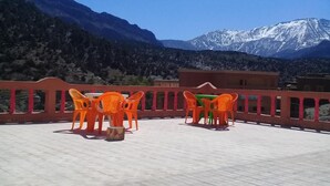 Une terrasse extérieure avec des chaises et des tables en plastique orange, une rambarde rouge et un paysage montagneux en arrière-plan.