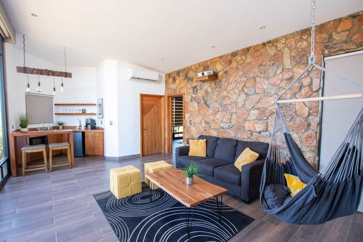 A modern living room with a stone wall, a hanging hammock, a sofa with cushions, a wooden coffee table, and a kitchen area with a bar stool and a plant.