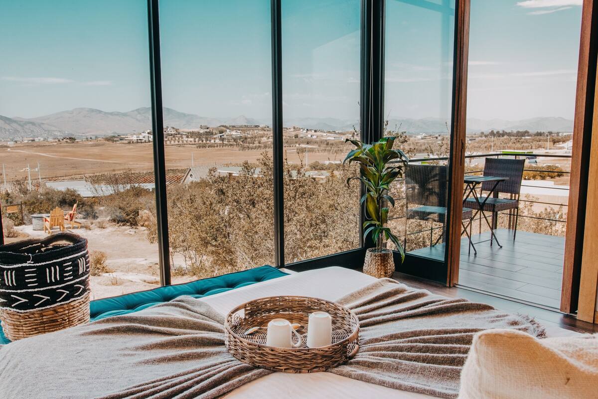 A bedroom with a large window, a wicker basket with candles, a plant, and a view of a landscape.