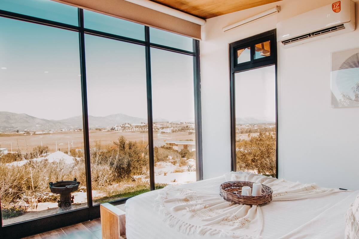 A bedroom with a large bed, wooden ceiling, and a view of a desert landscape through large windows.