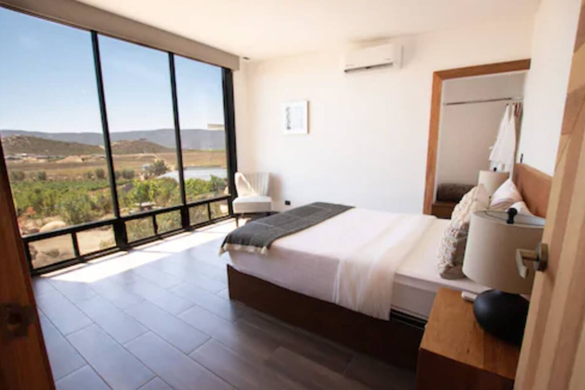 A bedroom with a large window, a bed, a bedside table, and a view of the outdoors.