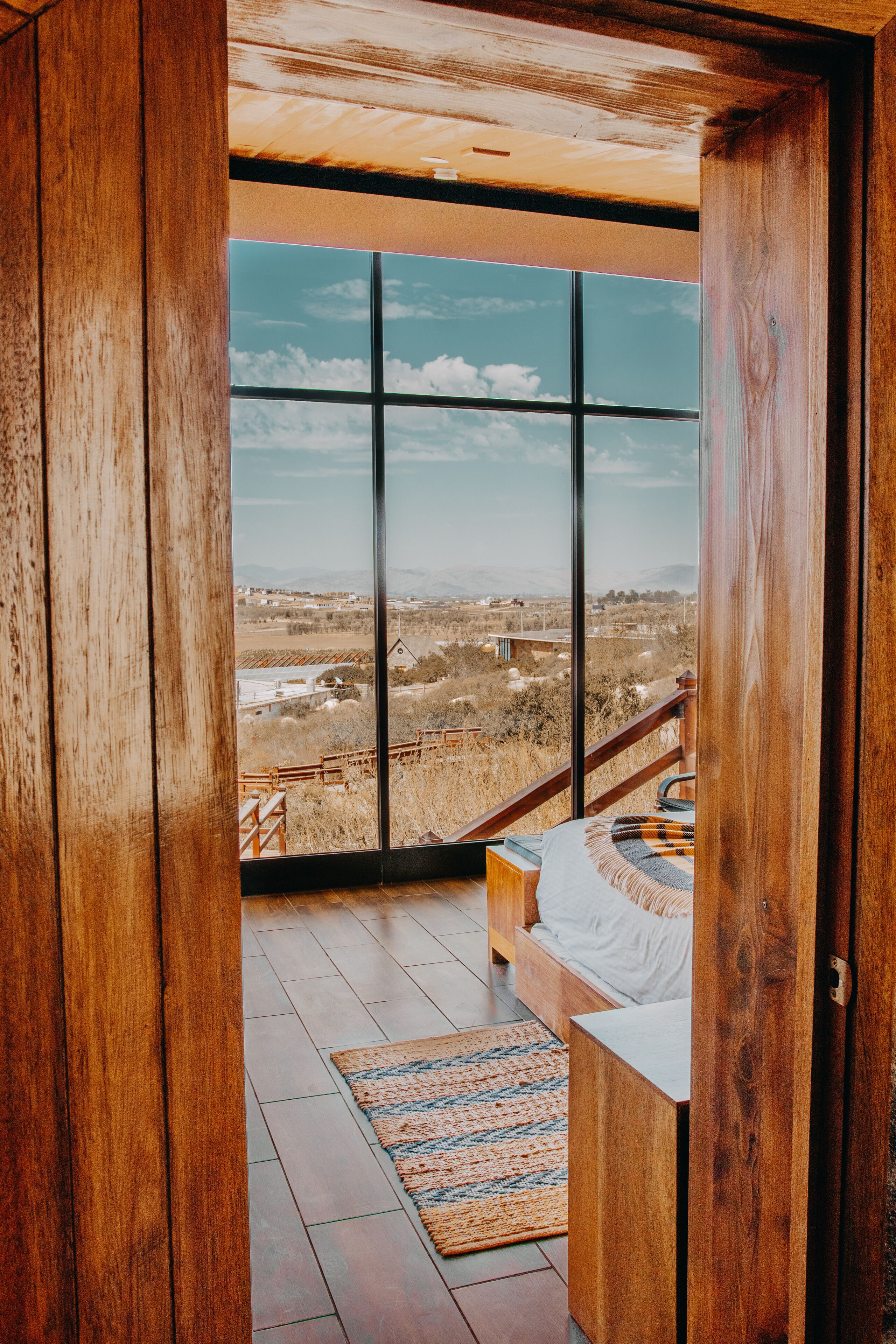 A room with a large window, wooden flooring, a bed, and a view of a landscape.