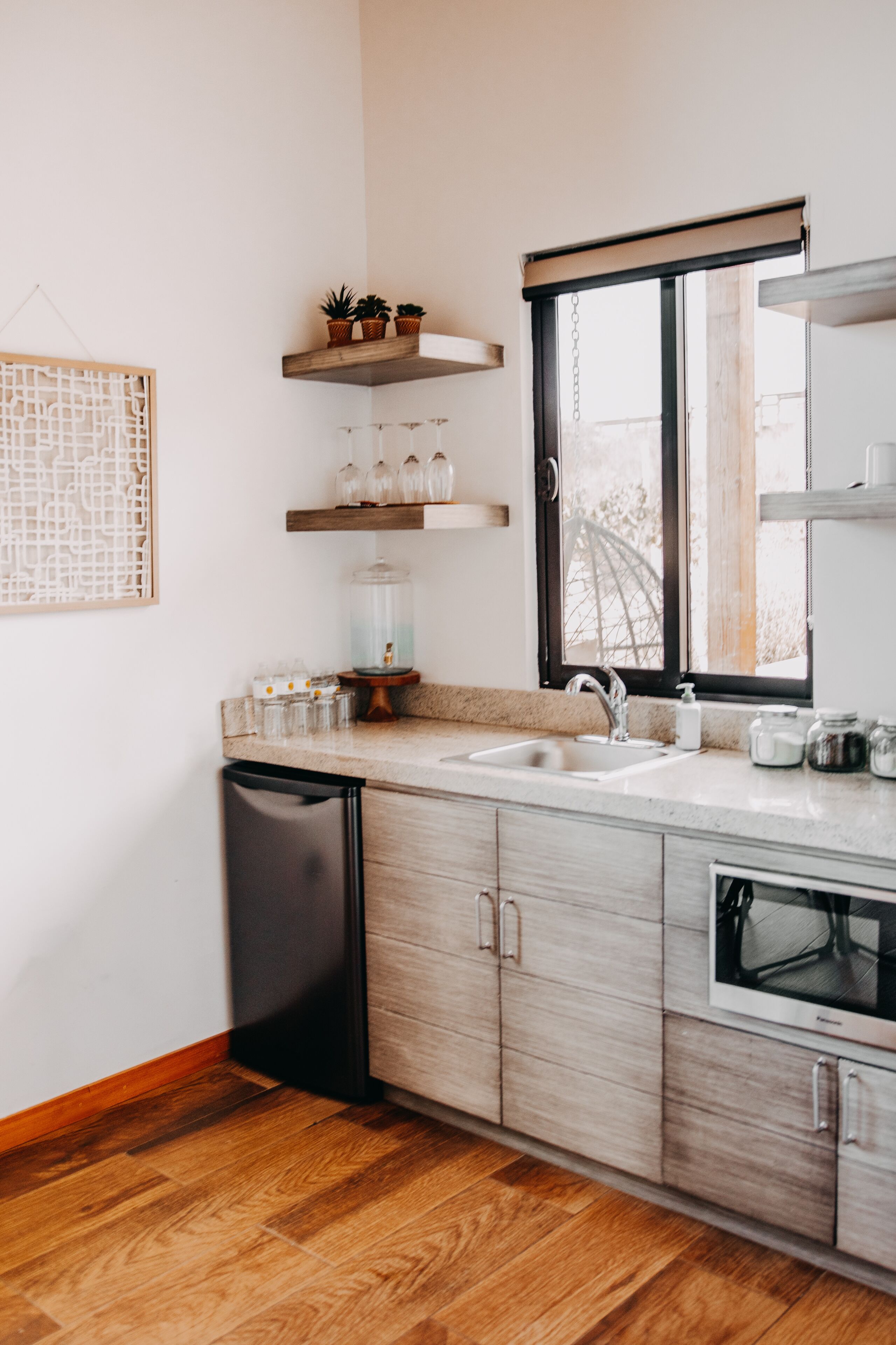 A modern kitchen with wooden cabinets, a black dishwasher, a window with a view, and a shelf with plants and jars.