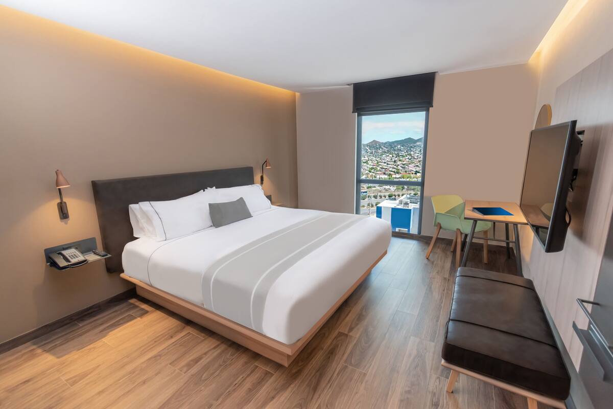 Ein modernes Hotelzimmer mit einem großen Bett, einem Schreibtisch, einem Sessel, einem Fernseher und Blick auf eine Stadtlandschaft.