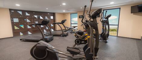 A gym with various cardio machines, a large window with a view, and a flat-screen TV mounted on the wall.