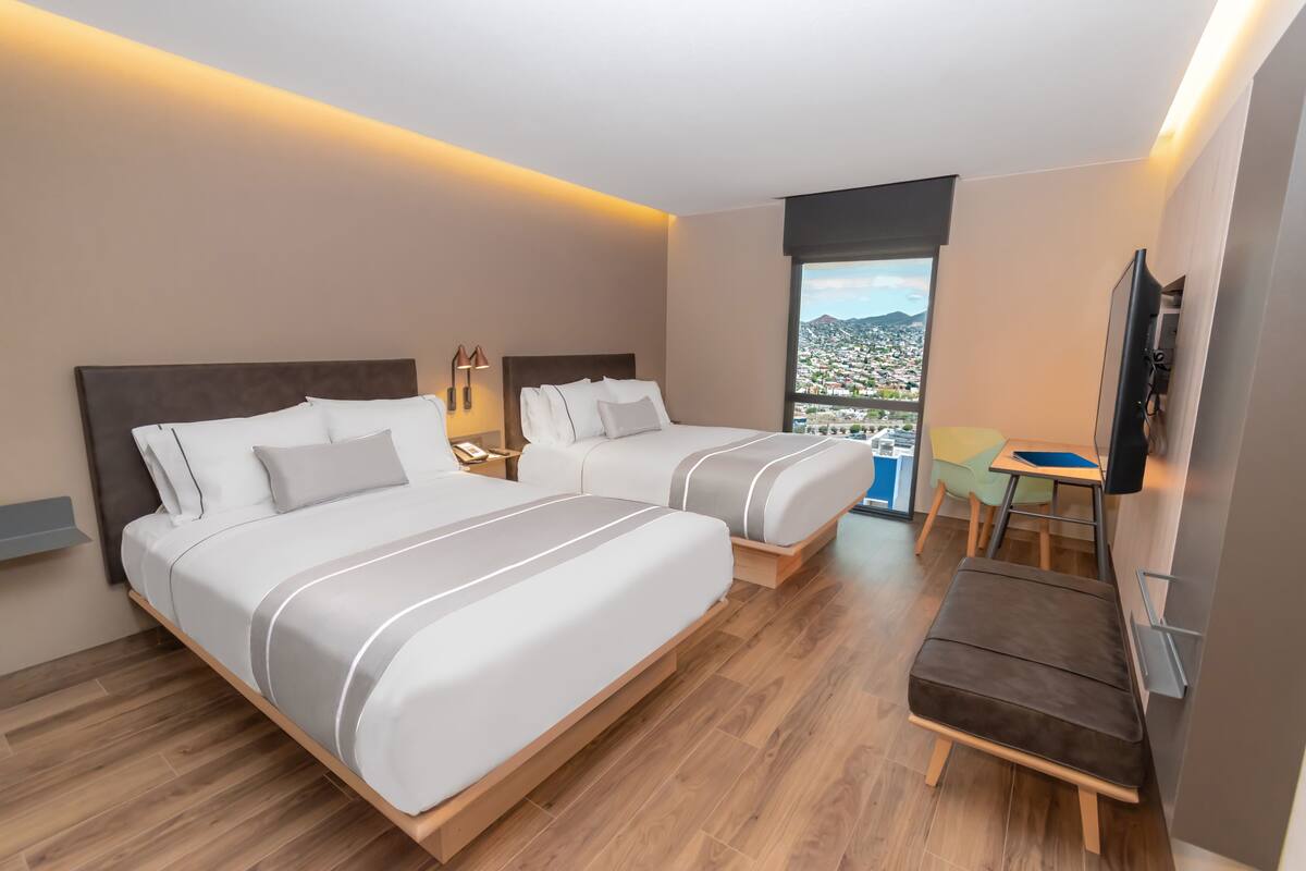 Ein Hotelzimmer mit zwei Betten, einem Schreibtisch, einem Stuhl, einem Fernseher und einem Balkon mit Blick auf die Stadt.