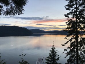 A tranquil lake with a wooden dock extending into the water, surrounded by dense forest and distant mountains. - —Priest Lake Waterfront Escape— (Coolin)