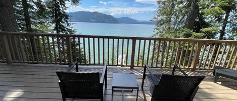 A wooden deck with two black wicker chairs and a small table, overlooking a lake with mountains in the distance.