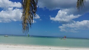 Une plage à l’eau turquoise et limpide, un rivage sablonneux et quelques bateaux à l’horizon.