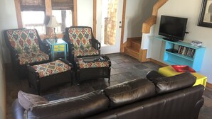 Ein Wohnzimmer mit einem braunen Ledersofa, zwei gemusterten Sesseln, einem kleinen Tisch mit einer Lampe, einem Fernseher auf einem Schrank und einem Bücherregal.