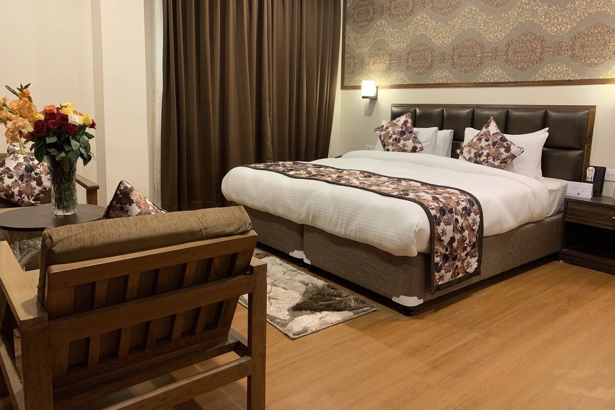 A hotel room with a large bed, a wooden chair, a table with a vase of flowers, and a patterned wall.