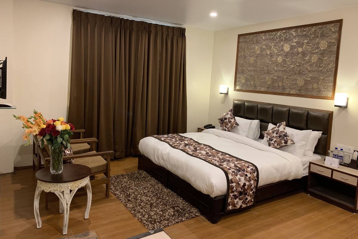 A hotel room with a large bed, a nightstand, a small table with a vase of flowers, and a large floral-patterned painting on the wall.