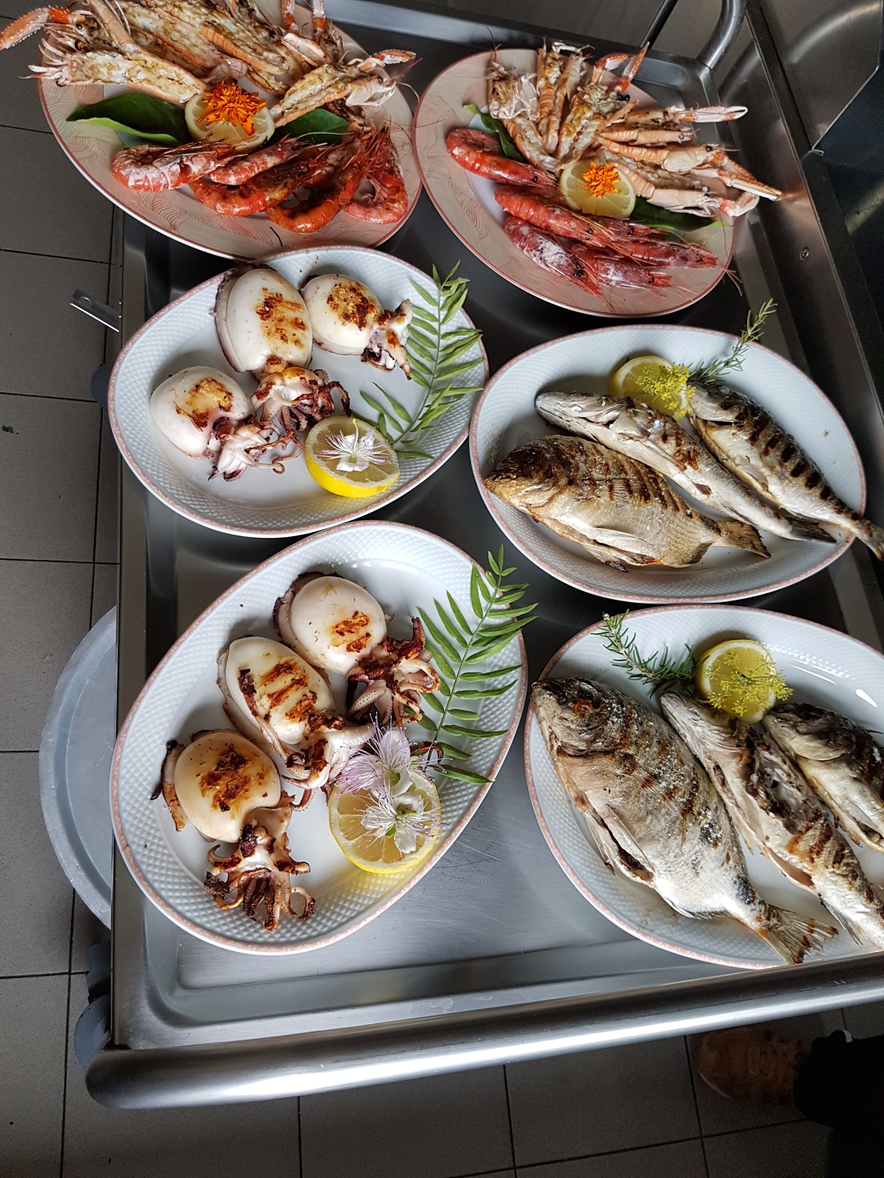 Eine Schale mit verschiedenen Meeresfrüchten, darunter gegrillter Fisch und Garnelen, serviert auf weißen Tellern mit Zitronenscheiben und Kräutern.