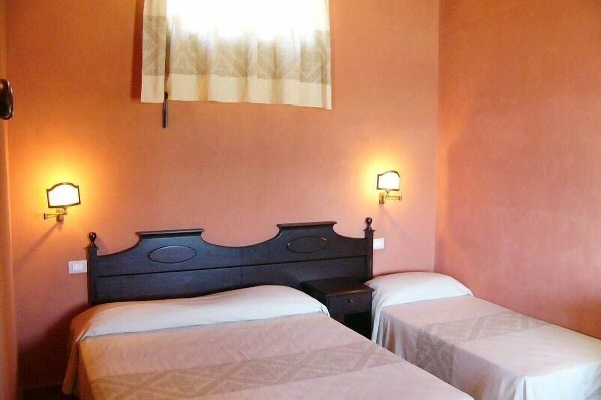 Habitación con dos camas individuales, un cabecero de madera oscura y dos mesitas de noche fijadas a la pared con lámparas.