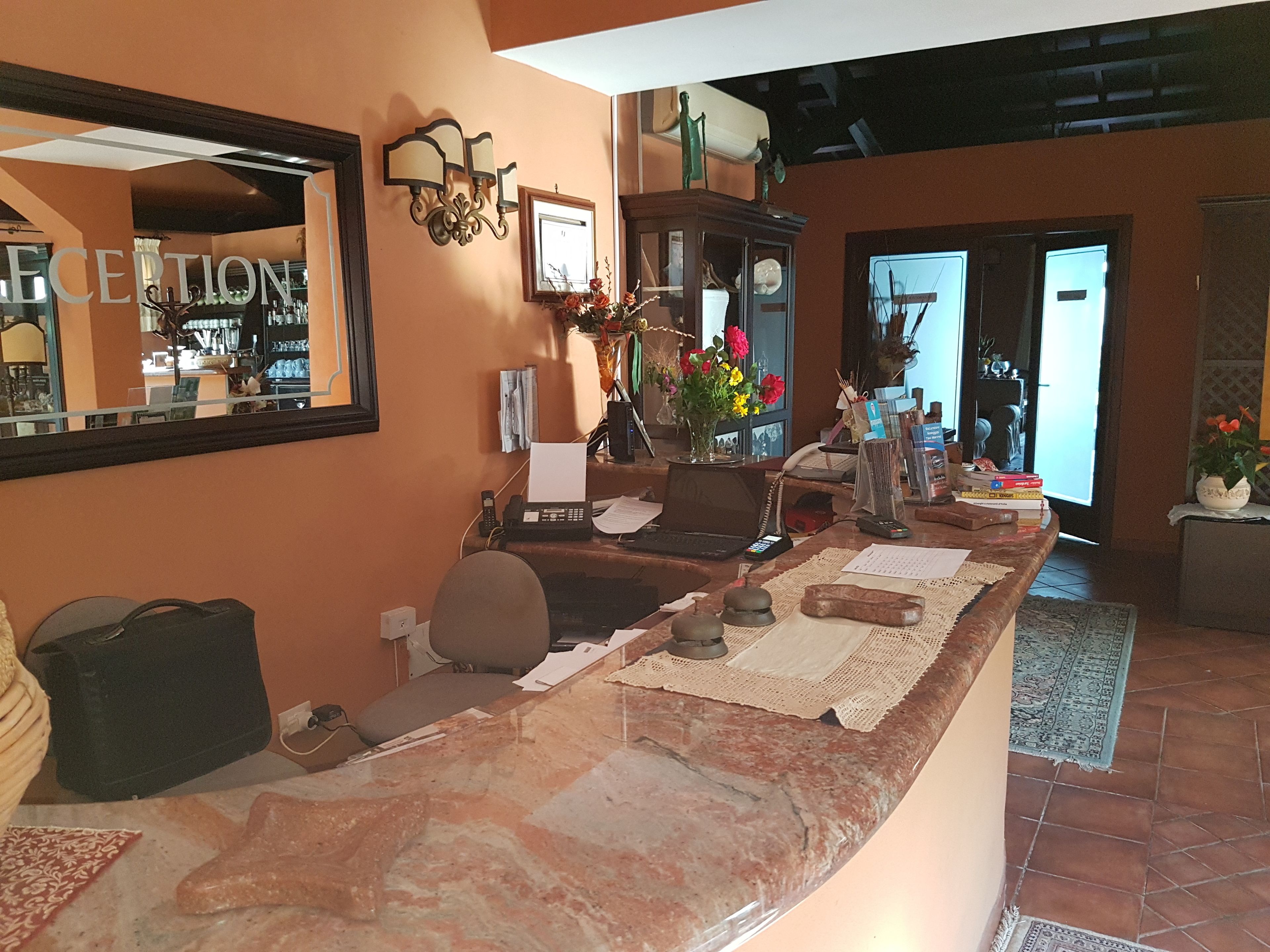 A reception area with a marble counter, a mirror, a telephone, a laptop, and a vase with flowers.