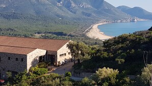 Edifici de pedra amb teulada vermella, carrer d'accés pavimentat i vista a zona costanera amb muntanyes i platja.